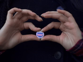 Two hands holding a pin featuring the silhouette of a cat's head in the colors of the trans flag.