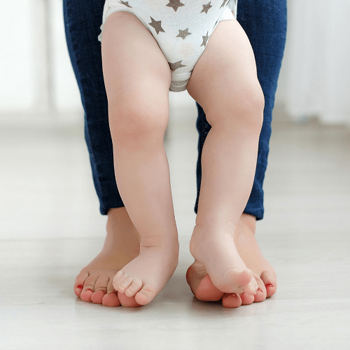 Barefoot person walking with a barefoot baby, the baby's feet resting on the adult's feet.