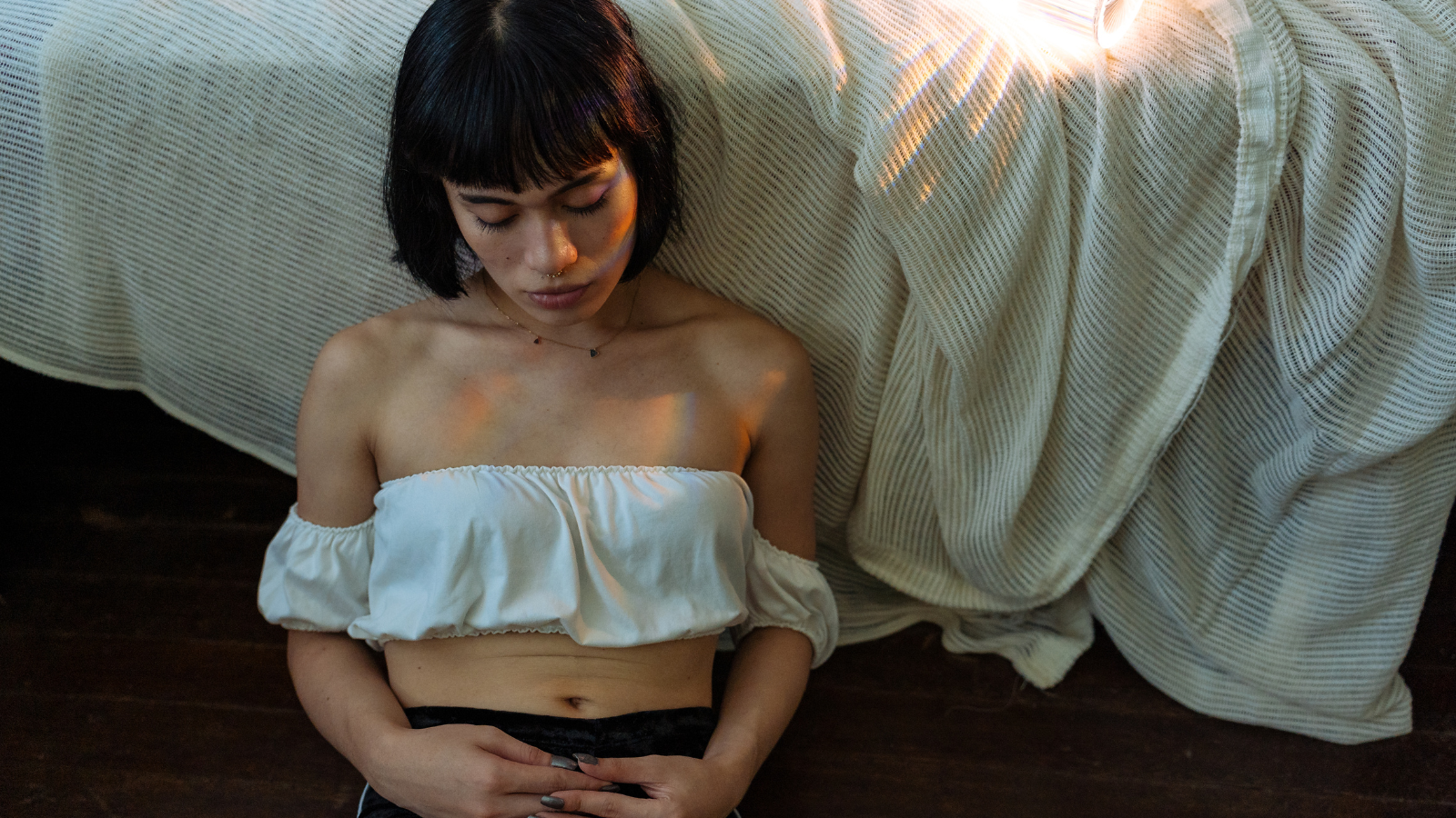 A trans woman sits against a bed looking down at her hands.